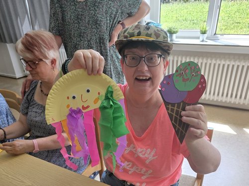 Eine strahlende Person mit Brille und Tarnmütze präsentiert zwei Bastelarbeiten: eine gelbe Papierqualle mit bunten Papierstreifen und ein mehrfarbiges Papier-Eis. Auf dem Holztisch liegen Bastelutensilien wie Kleber und Stifte.