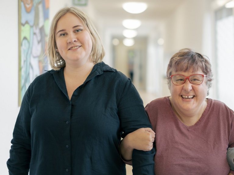 Eine Mitarbeiterin der Behindertenhilfe Haus Nettetal geht mit einer behinderten Bewohnerin Arm in Arm über den Flur