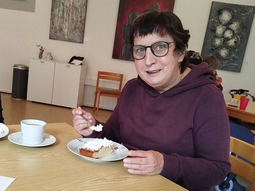 Eine Frau mit Brille und lila Kapuzenpullover sitzt an einem Holztisch und isst mit einer Gabel ein Stück Kuchen. Auf dem Tisch stehen eine weiße Tasse und ein blauer Becher. Im Hintergrund hängen Bilder an der Wand.