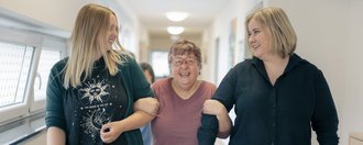 Zwei Mitarbeiterinnen gehen mit einer behinderten Frau fröhlich über den Flur