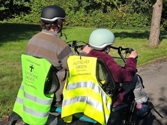 Zwei Personen fahren von hinten gesehen auf einem Tandemfahrrad. Beide tragen Helme und leuchtend gelbe Warnwesten mit dem Logo „Deutscher Orden“. Im Hintergrund sind Bäume und Gras zu sehen.
