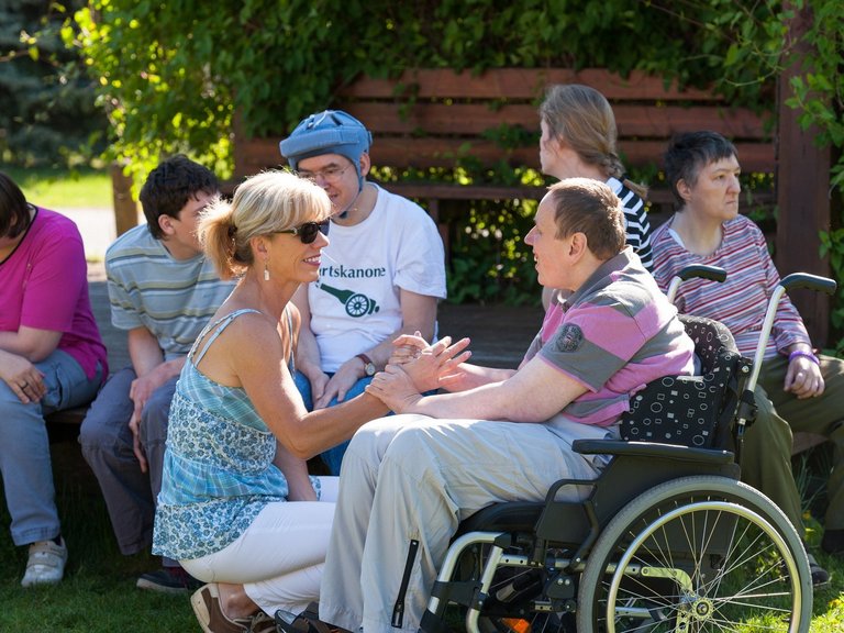 Eine Pflegerin kniet vor einem Mann, der im Rollstuhl sitzt - beide lachen