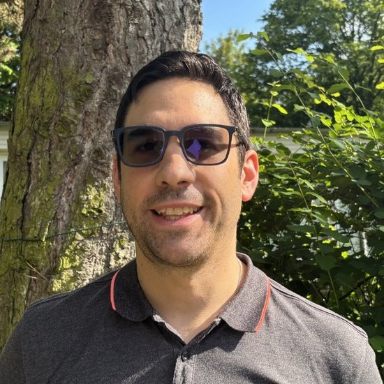Ein lächelnder Mann mit dunklen Haaren und Sonnenbrille steht im Freien vor einem Baum. Er trägt ein graues Poloshirt mit roten Kragen- und Ärmelbündchen.