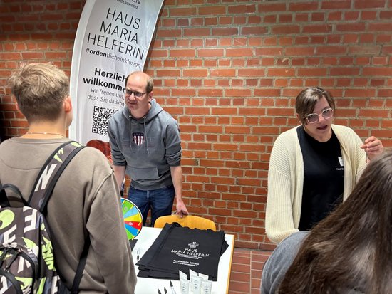 Bei einem Informationsstand vor einer Backsteinwand stehen ein Mann und eine Frau hinter einem Tisch mit einem schwarzen T-Shirt. Ein Banner im Hintergrund zeigt „Haus Maria Helferin“. Ein junger Besucher mit Rucksack steht ihnen gegenüber.