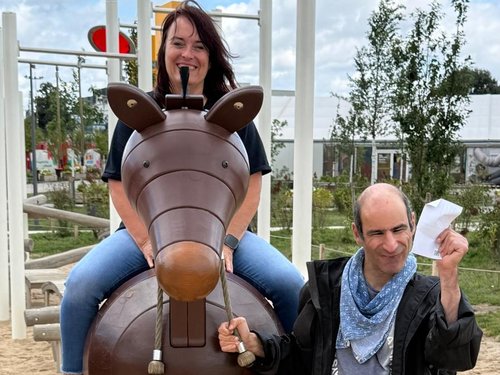 Eine Frau sitzt lächelnd auf einem braunen, tierförmigen Spielgerät. Ein Mann steht daneben und hält ein weißes Blatt Papier hoch. Beide wirken fröhlich auf einem sandigen Spielplatz.