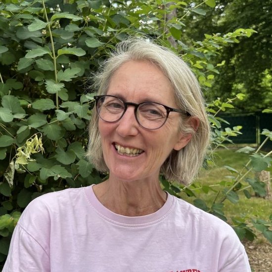 Eine lächelnde Frau mit kurzen grauen Haaren und Brille steht vor einem großen grünen Busch. Sie trägt ein hellrosa T-Shirt mit einem roten Erdbeer-Aufdruck.