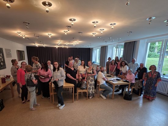 Eine große Gruppe von Menschen steht in einem hellen Raum und posiert für ein Gruppenfoto. Im Hintergrund sind Fenster mit Blick ins Grüne sowie Tische mit verschiedenen Gegenständen zu sehen.