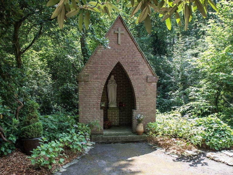 Madonnenstatue in einer kleinen Kapelle in einem Park umringt von vielen Pflanzen und Bäumen