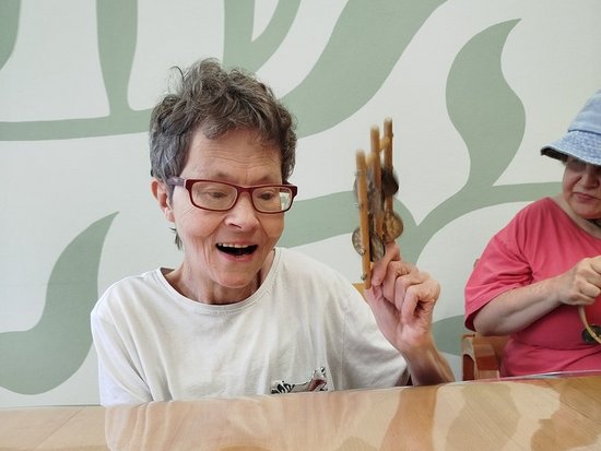 Eine Frau mit kurzen Haaren und Brille hält ein kleines, braunes Objekt in die Höhe und lacht begeistert. Sie sitzt an einem Holztisch, während rechts eine weitere Person teilweise im Hintergrund zu sehen ist.