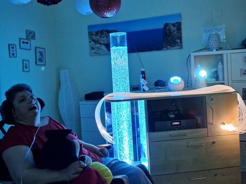 Eine Frau im Rollstuhl sitzt in einem blau beleuchteten Raum und lacht fröhlich. Sie trägt Kopfhörer und hält ein schwarzes Plüschtier. Eine leuchtende Blasenlampe und hängende Papierlaternen sorgen für stimmungsvolles Licht. Auf einem Schrank steht eine Ringlampe.
