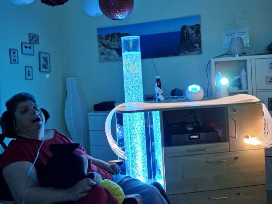 Eine Frau im Rollstuhl sitzt in einem blau beleuchteten Raum und lacht fröhlich. Sie trägt Kopfhörer und hält ein schwarzes Plüschtier. Eine leuchtende Blasenlampe und hängende Papierlaternen sorgen für stimmungsvolles Licht. Auf einem Schrank steht eine Ringlampe.