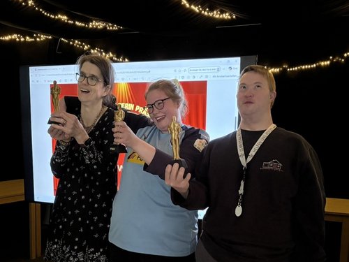 Drei Personen stehen auf einer Bühne vor einer Leinwand und machen ein Selfie. Über ihnen hängen Lichterketten. Zwei der Personen tragen Medaillen um den Hals.