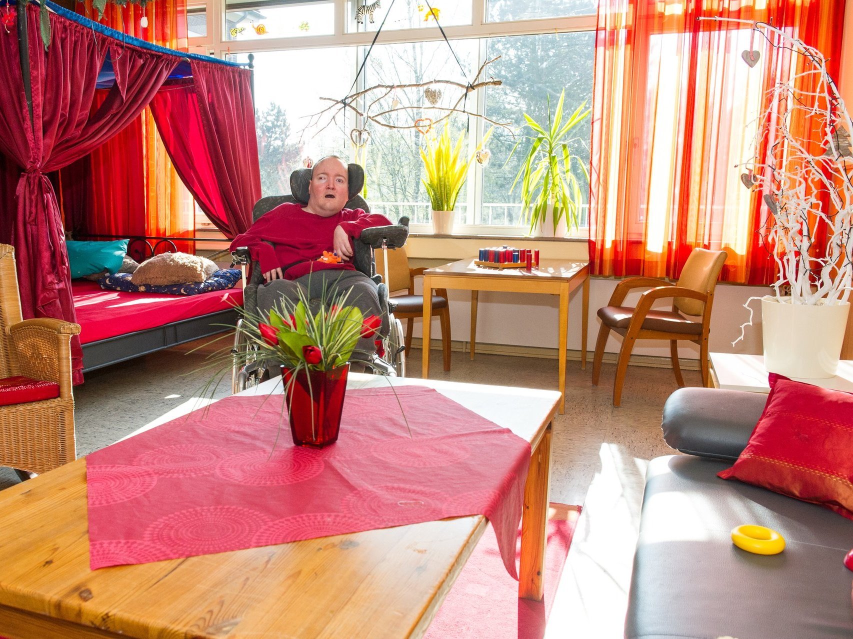 Ein Mann im Rollstuhl in einem gemütliches Zimmer mit Bett, Sessel, Regal und orangen Vorhängen