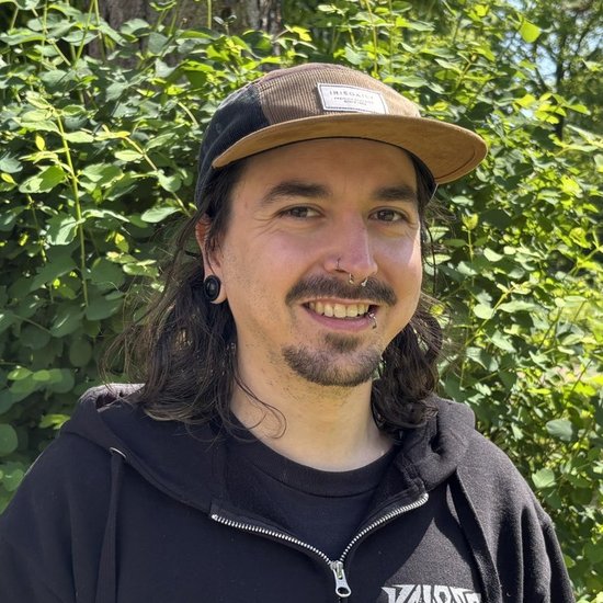 Ein lächelnder Mann mit langen dunklen Haaren, einem Bart und einer Kappe steht im Freien vor grünen Büschen. Er trägt einen schwarzen Kapuzenpullover mit weißem Logo.