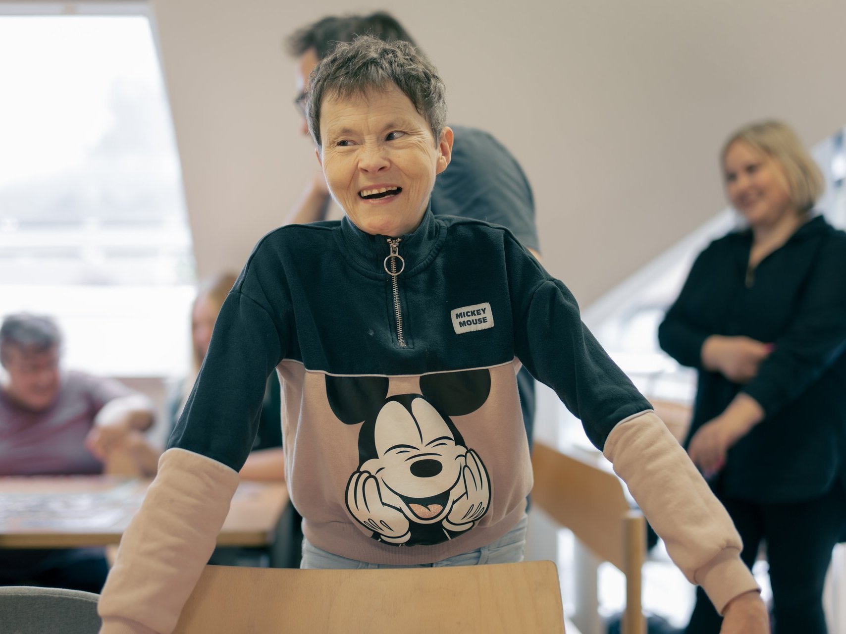 Eine behinderte Frau mit kurzen Haaren steht hinter einem Stuhl und lacht in die Kamera