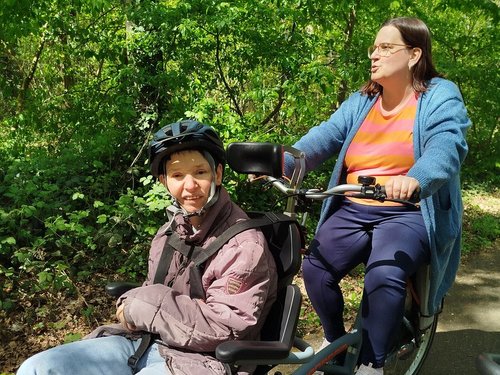 Zwei Frauen fahren gemeinsam auf einem Tandemfahrrad durch einen sonnigen Wald. Die Frau vorne sitzt in einem speziellen Sitz mit Helm, während die Frau hinten in die Pedale tritt. Sie genießen die Fahrt im Grünen.