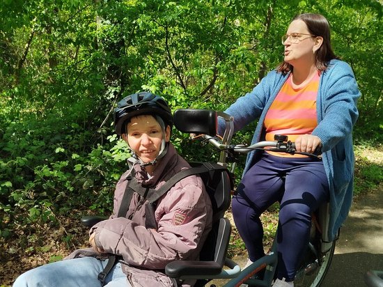 Zwei Frauen fahren gemeinsam auf einem Tandemfahrrad durch einen sonnigen Wald. Die Frau vorne sitzt in einem speziellen Sitz mit Helm, während die Frau hinten in die Pedale tritt. Sie genießen die Fahrt im Grünen.