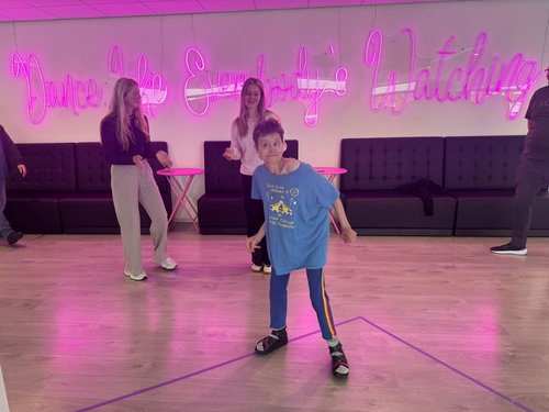 Ein Junge in einem blauen T-Shirt tanzt in einem Tanzstudio. Im Hintergrund stehen Erwachsene. An der Wand leuchtet eine große rosa Neon-Schrift: "Dance Like Everybody's Watching".