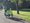 Zwei Personen fahren auf einem Fahrrad auf einem asphaltierten Weg durch einen Park. Beide tragen leuchtend gelbe Warnwesten und sind von hinten zu sehen.