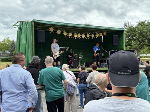 Eine Band spielt auf einer kleinen, grünen Freiluftbühne, die mit gelben Sonnen-Schablonen geschmückt ist. Ein Publikum steht im Vordergrund und hört dem Auftritt zu.