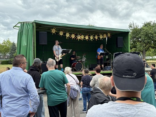 Eine Band spielt auf einer kleinen, grünen Freiluftbühne, die mit gelben Sonnen-Schablonen geschmückt ist. Ein Publikum steht im Vordergrund und hört dem Auftritt zu.