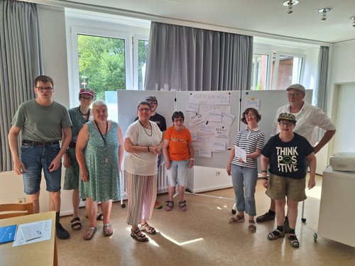 Eine Gruppe von neun Menschen steht in einem hellen Raum vor Whiteboards mit Notizen und Skizzen. Die Personen tragen lässige Sommerkleidung und posieren lächelnd für ein Gruppenfoto.